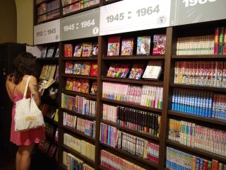 Historical manga (lower) A large bookcase full of manga, with a woman visible to the left reading one of them. Along the top are signs with year-ranges.